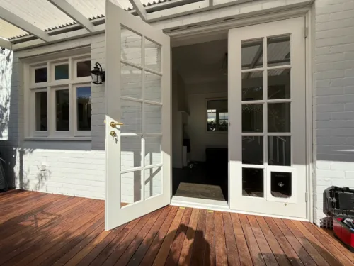 A pet door fitted in the lower panel of white French doors that open onto a timber deck