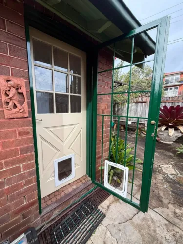 A green security screen door held open to show the second pet door fitted in the cream door behind it