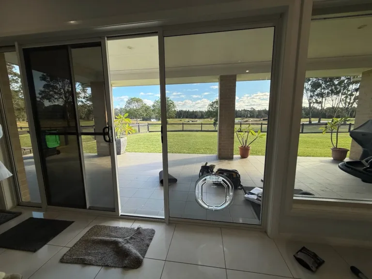 A pet door fitted in a sliding glass door, with a dog resting inside and a grassy field outside