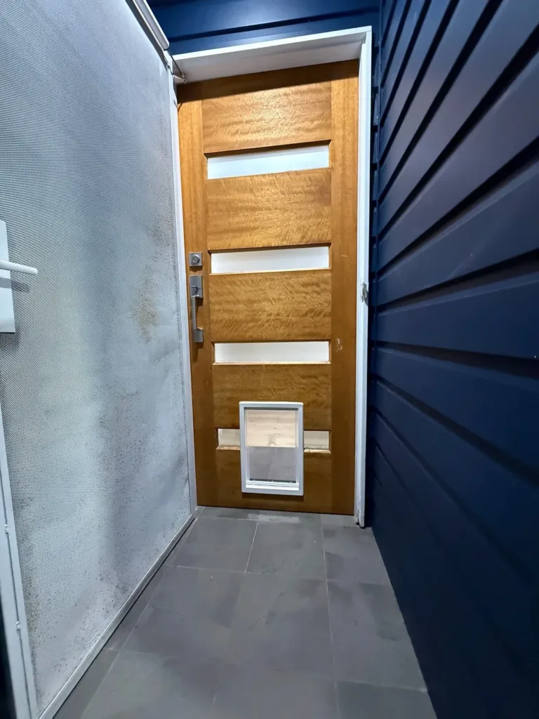 A white pet door at the base of a timber door with vertical glass panels, on a blue weatherboard wall