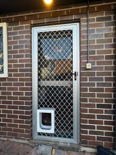 A white pet door at the base of a diamond-mesh security door on a brick home